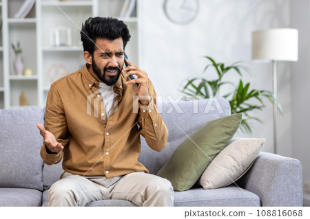 Angry angry man talking on the phone at home, dissatisfied customer sitting on the sofa in the living room yelling at the interlocutor. 108816068