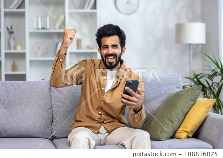 Portrait of winner at home, happy happy man looking at camera, holding phone, holding hand up in triumph gesture, satisfied with achievement results, sitting on sofa in living room. Portrait of winner at home, happy happy man looking at camera, holding phone, holding hand up in triumph gesture, satisfied with achievement results, sitting on sofa in living room. 108816075