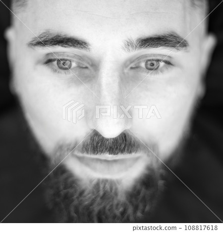 Black and white portrait of a bearded man closeup. A young man with a beard in a black cape sits on a chair in a barbershop. The client is waiting for a haircut of the beard. Black and white portrait of a bearded man closeup. A young man with a beard in a black cape sits on a chair in a barbershop. The client is waiting for a haircut of the beard. 108817618