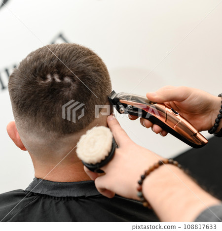 Barber shaves the contour of the oval line with a clipper on the client head. A man with a beard gets a haircut in a barbershop chair. 108817633