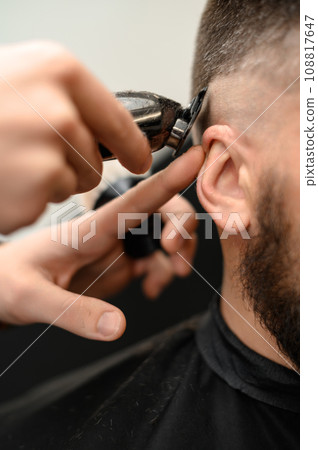 Barber shaves the contour of the oval line with a clipper on the client head. A man with a beard gets a haircut in a barbershop chair. 108817647