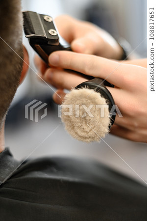 Barber shaves the contour of the oval line with a clipper on the client head. A man with a beard gets a haircut in a barbershop chair. Barber shaves the contour of the oval line with a clipper on the client head. A man with a beard gets a haircut in a barbershop chair. 108817651