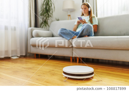 A woman in headphones sits on couch while a robotic vacuum cleaner does cleaning 108817899