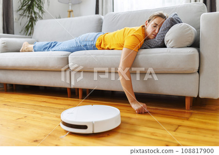 A woman sleeping on sofa while robot vacuum quietly takes care of the cleaning 108817900