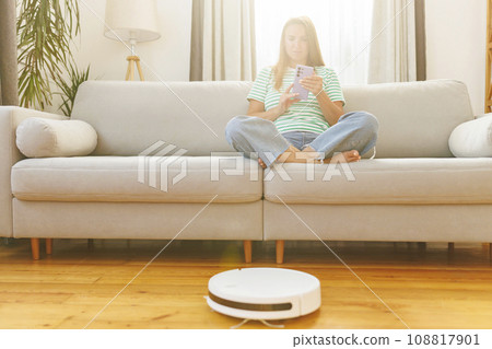 A woman in headphones sits on couch while a robotic vacuum cleaner does cleaning 108817901