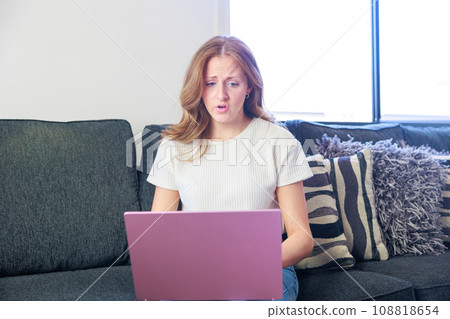 A white woman sitting on a sofa and using a computer (surprised/sad) 108818654