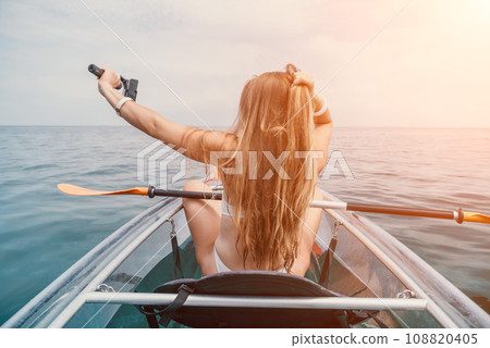 Woman in kayak back view. Happy young woman with long hair floating in transparent kayak on the crystal clear sea. Summer holiday vacation and cheerful female people relaxing having fun on the boat Woman in kayak back view. Happy young woman with long hair floating in transparent kayak on the crystal clear sea. Summer holiday vacation and cheerful female people relaxing having fun on the boat 108820405