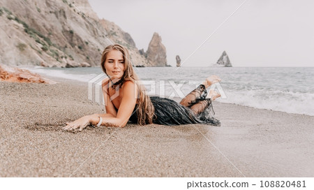 Woman summer travel sea. Happy tourist in black dress enjoy taking picture outdoors for memories. Woman traveler posing on sea beach surrounded by volcanic mountains, sharing travel adventure journey Woman summer travel sea. Happy tourist in black dress enjoy taking picture outdoors for memories. Woman traveler posing on sea beach surrounded by volcanic mountains, sharing travel adventure journey 108820481