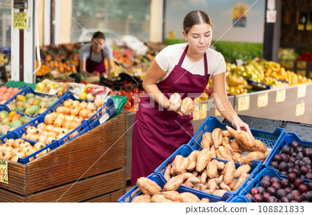Girl seller of fruit and vegetable store works near showcase with garden-stuff, sorting yam Girl seller of fruit and vegetable store works near showcase with garden-stuff, sorting yam 108821833