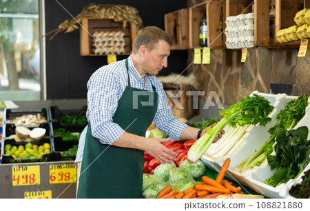 Man seller of fruit and vegetable store works near showcase, sorting greenery celery Man seller of fruit and vegetable store works near showcase, sorting greenery celery 108821880