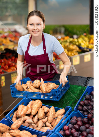 Smiling saleswoman lying out boxes of sweet potatoes in farmers market Smiling saleswoman lying out boxes of sweet potatoes in farmers market 108821926