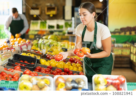 Girl seller of garden stuff store works near showcase, sorting tomato, checking goods Girl seller of garden stuff store works near showcase, sorting tomato, checking goods 108821941