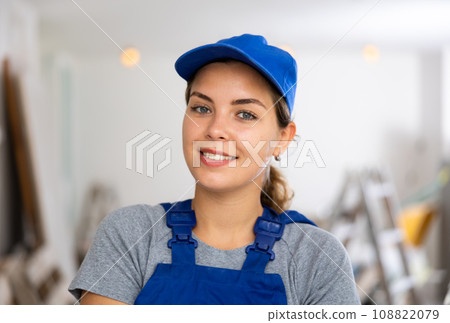 Portrait of contented young female builder in blue overalls during the repair of cottage Portrait of contented young female builder in blue overalls during the repair of cottage 108822079