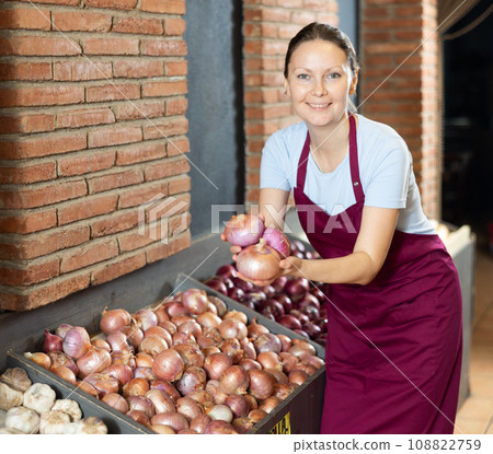 Woman seller of fruit and vegetable store works near showcase with garden-stuff, sorting onion 108822759