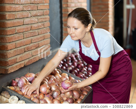 Woman seller of fruit and vegetable store works near showcase with garden-stuff, sorting onion Woman seller of fruit and vegetable store works near showcase with garden-stuff, sorting onion 108822869