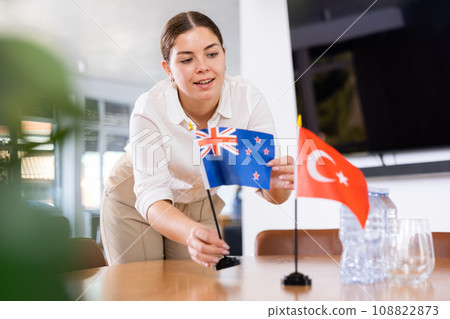 Young female assistant putting up turkish and new zealand flags on table for international negotiations Young female assistant putting up turkish and new zealand flags on table for international negotiations 108822873