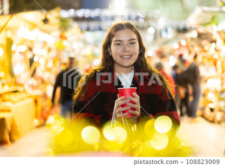 Girl with paper cup of tea walking at street Christmas fair 108823009