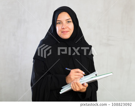 gleeful girl in hijab holds several notebooks and writes important note in her diary gleeful girl in hijab holds several notebooks and writes important note in her diary 108823092