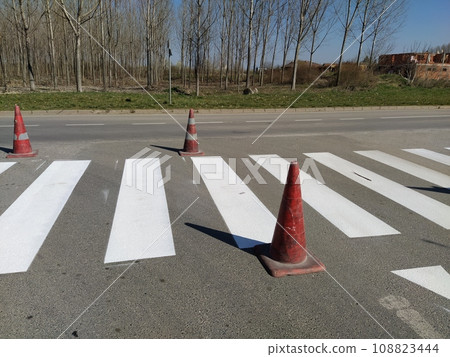 Painting pedestrian crossings with new paint. Road warning plastic orange cone. White stripes on the pavement. The pedestrian crossing is marked with special road signs or markings 108823444