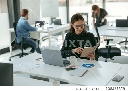 Smiling female entrepreneur holding note pad while working on laptop sitting in modern coworking 108825859
