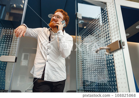 Bearded businessman in eyeglasses is talking phone standing in modern coworking space Bearded businessman in eyeglasses is talking phone standing in modern coworking space 108825868