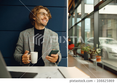 Cheerfull male manager use mobile phone and drink coffee while sitting in cafe near window Cheerfull male manager use mobile phone and drink coffee while sitting in cafe near window 108825869