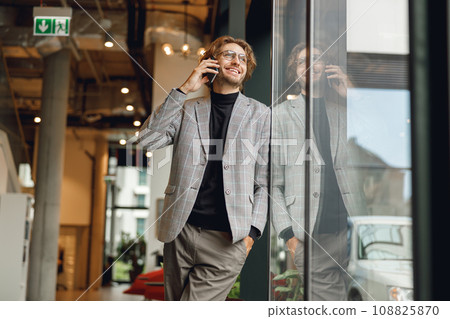 Smiling businessman talking by mobile phone standing in coworking near window and looks away Smiling businessman talking by mobile phone standing in coworking near window and looks away 108825870