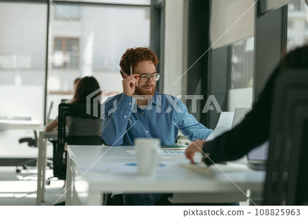 Smiling male sales manager in eyeglasses working on laptop sitting in office during working day Smiling male sales manager in eyeglasses working on laptop sitting in office during working day 108825963