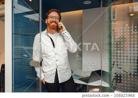 Bearded male freelancer in eyeglasses is talking phone standing in modern coworking space 108825968