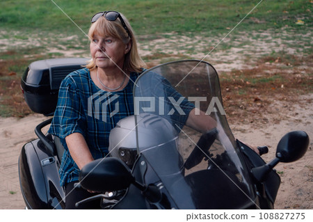 Strong independent mature blonde looks seriously to side while sitting on motorcycle on autumn. 108827275