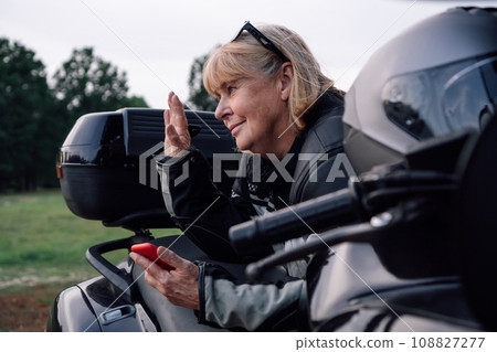 Mature blonde on black motorcycle greets someone, holding mobile phone in nature during stop. 108827277