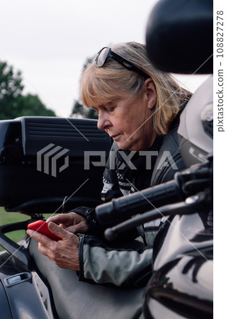 Communication in travel. Mature blonde on motorcycle, holding mobile phone in nature during stop. 108827278