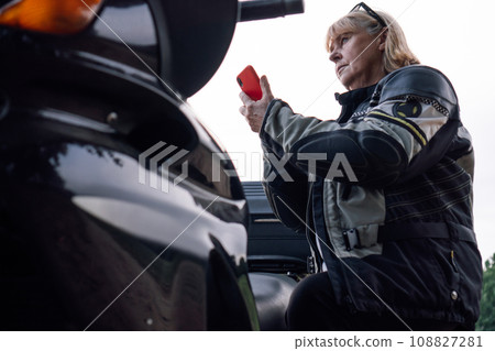 Bottom view of confident, serious mature blonde biker talking on red phone near black motorcycle. Bottom view of confident, serious mature blonde biker talking on red phone near black motorcycle. 108827281