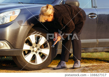 Unrecognizable blonde woman is leaning over car and changing broken tire on autumn day in nature. Unrecognizable blonde woman is leaning over car and changing broken tire on autumn day in nature. 108827317
