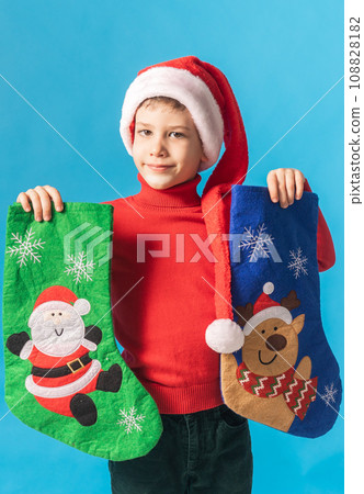 Child boy in red sweater and Santa hat with Christmas socks on blue background. Xmas and holidays concept. Generation alpha and gen alpha children. 108828182