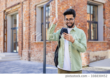 Young indian student received online win notification message phone, successful entrance exam results, man with backpack walking outside university campus, celebrating successful achievement result. 108830452