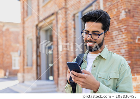 Portrait of young successful student outside university campus, man smiling and using app on phone, typing message and browsing social media, indian man holding phone with backpack. 108830455