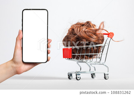 A female's hand holds a smartphone with a white screen. A shopping cart filled with brown hair falling down. White background. Mock up. The concept of buying and selling hair A female's hand holds a smartphone with a white screen. A shopping cart filled with brown hair falling down. White background. Mock up. The concept of buying and selling hair 108831262