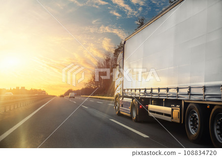 Tail view white blank modern delivery big shipment cargo commercial semi trailer truck moving motorway road city urban suburb. Business distribution logistics service. Lorry driving highway sunset Tail view white blank modern delivery big shipment cargo commercial semi trailer truck moving motorway road city urban suburb. Business distribution logistics service. Lorry driving highway sunset 108834720