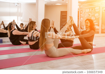 A group of six athletic women doing pilates or yoga on pink mats in front of a window in a beige loft studio interior. Teamwork, good mood and healthy lifestyle concept. 108837349