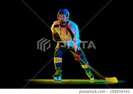 Young girl, hockey player in uniform and helmet training, standing with stick against black studio background in neon light 108838342