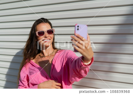 Young beautiful brunette in a pink shirt, neck jewelry, necklace, trendy sunglasses on the background of a light garage fence, takes a selfie on the phone, laughs, smiles Young beautiful brunette in a pink shirt, neck jewelry, necklace, trendy sunglasses on the background of a light garage fence, takes a selfie on the phone, laughs, smiles 108839368