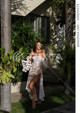 Young beautiful woman in a romantic dress with a floral print, sunglasses and a pearl necklace bracelet, against the backdrop of tropical leaves Young beautiful woman in a romantic dress with a floral print, sunglasses and a pearl necklace bracelet, against the backdrop of tropical leaves 108839374
