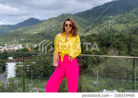Stylish fit fashion women in bright pink wide leg pants and yellow shirt holding bag trendy sunglasses posing at rooftop terrace tropical view outdoor natural day light Stylish fit fashion women in bright pink wide leg pants and yellow shirt holding bag trendy sunglasses posing at rooftop terrace tropical view outdoor natural day light 108839400