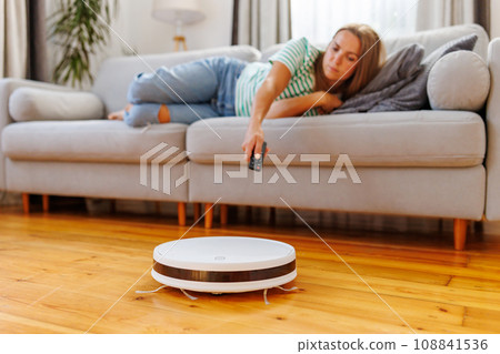 Robot vacuum cleaner cleaning a room while a woman relaxes on the sofa Robot vacuum cleaner cleaning a room while a woman relaxes on the sofa 108841536