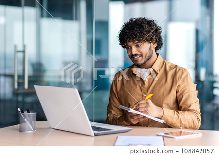 Man studying online remotely, businessman improving qualifications at the workplace inside office, writing down data in a notebook, watching a video course, satisfied with high results, smiling. 108842057