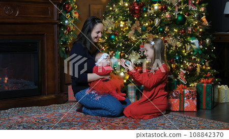 Mother with children having fun near Xmas tree, little girl playing with cute baby in Santas costume at home Mother with children having fun near Xmas tree, little girl playing with cute baby in Santas costume at home 108842370