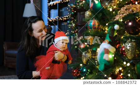 Smiling happy mother playing with cute baby daughter in Santas costume near Xmas tree, showing her Christmas decorations and balls Smiling happy mother playing with cute baby daughter in Santas costume near Xmas tree, showing her Christmas decorations and balls 108842372