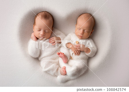 Tiny newborn twin boys in white bodysuits on a white background. Newborn twins sleep next to their brother on the background of the heart. Two newborn twin boys hugging each other. 108842574