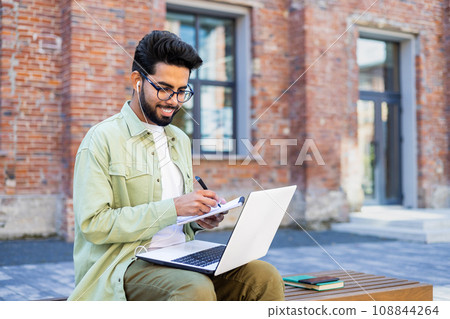 Young man studying remotely sitting on a bench outside a university building, a student in headphones watching a video online course, talking using a video call, writing down data in a notebook. Young man studying remotely sitting on a bench outside a university building, a student in headphones watching a video online course, talking using a video call, writing down data in a notebook. 108844264